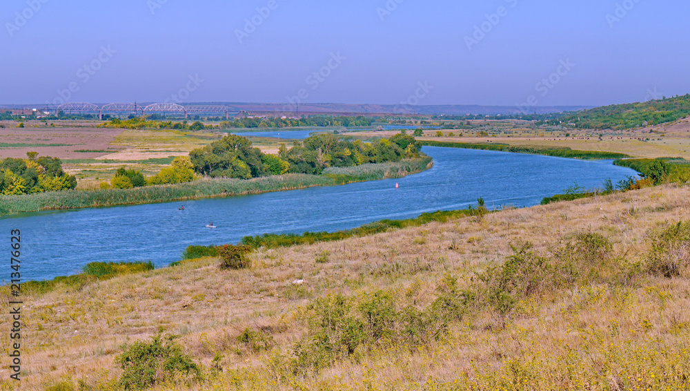 A wide river flowing between fields and low slopes covered with dry grass with peacefully grazing cows in the meadow.