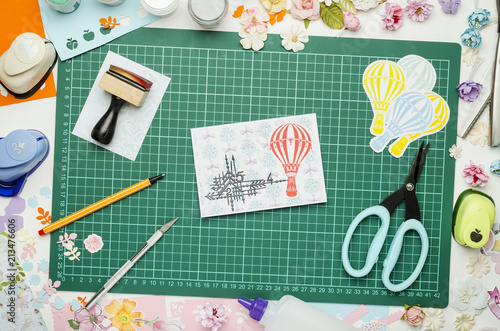 Multi-colored paper, homemade postcard and scrapbooking tools and materials on green mat for cutting, top view, no hands
