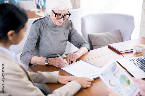 Senior woman wearing eyeglases writing testament with notary agent sitting near by her photo