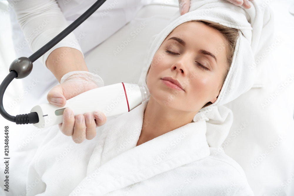 Pleased woman having procedure with gadget in beauty salon. Worker making it with digital device Pleased woman having procedure with gadget in beauty salon. Worker making it with digital device
