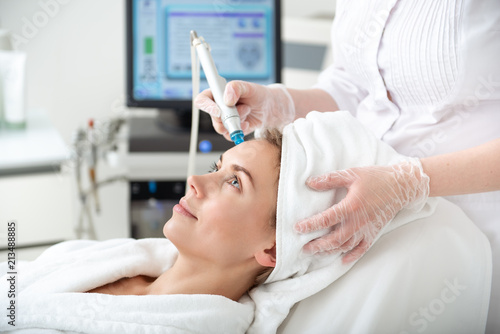 Side view smiling lady getting therapy for cleaning face. Cosmetologist doing it with special appliance in hands