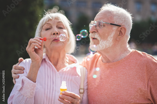 Back to childhood. Delighted senior man and woman standing on street and blowing bubbles with joy. Mature man is hugging wife while she is holding bottle with soap water