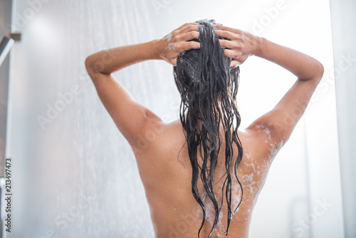 Slim young female enjoying shower while washing hair with shampoo. She turning back to camera