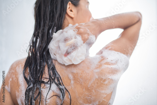 Youth female with wet hair washing body while holding fluffy sponge in hand. Hygiene concept