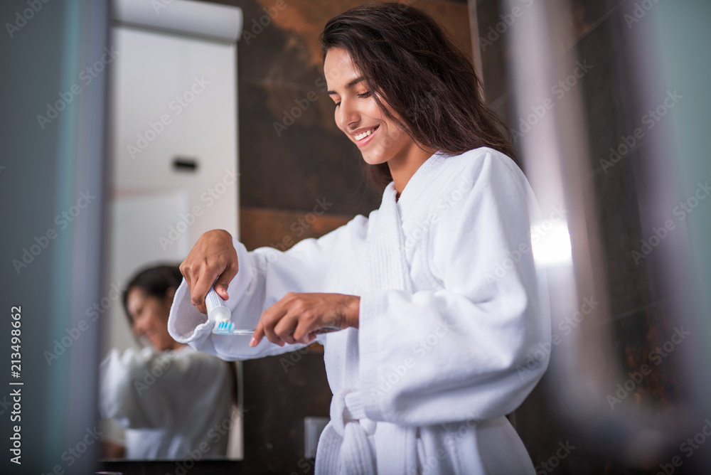 Fototapeta premium Portrait of happy girl squeezing toothpaste on toothbrush. She wearing bathrobe. She gesticulating hands