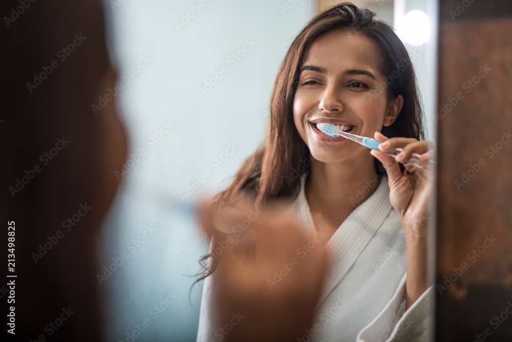 Fototapeta premium Portrait of beaming pretty woman brushing teeth while looking at mirror