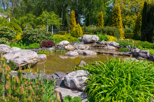 decorative pond surrounded by boulders, flowers, green bushes, deciduous and coniferous trees