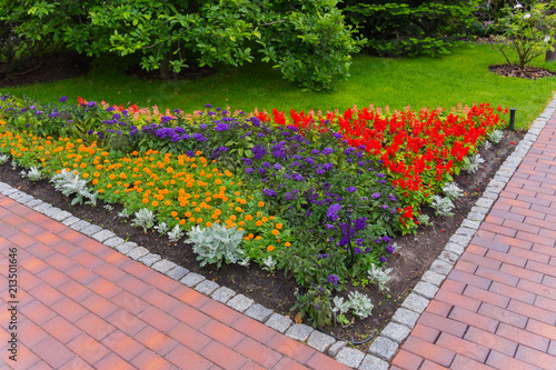 corner flowerbed with bright orange, blue and red flowers at the intersection of tiled garden paths against the backdrop of dense green bushes
