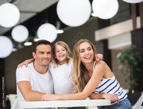 Waist up portrait of joyful couple sitting with child in bar at table. Small girl is pointing with finger while smiling woman and man are looking sideways. They are having fun while waiting for order