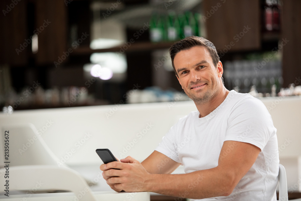 Waist up portrait of joyful man sitting at table in cafe. He is holding his phone in hands and surfing net with smile Waist up portrait of joyful man sitting at table in cafe. He is holding his phone in hands and surfing net with smile