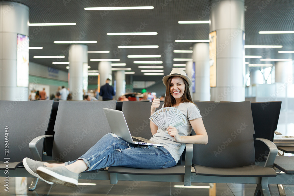 Young traveler tourist woman with laptop, hold bundle of dollars, cash money showing thumb up blinking wait in lobby hall at airport. Passenger traveling abroad on weekend getaway. Air flight concept.