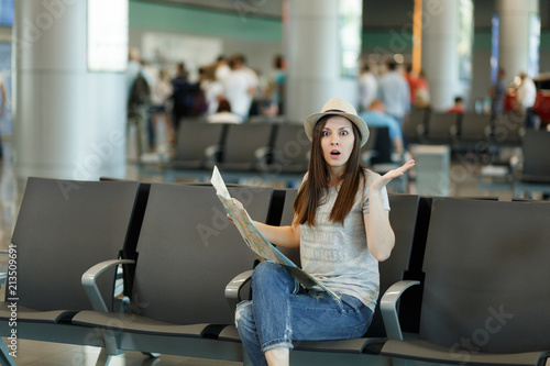 Young shocked traveler tourist woman holding paper map, search route, spreading hands wait in lobby hall at airport. Passenger traveling abroad on weekends getaway. Air travel, flight journey concept.