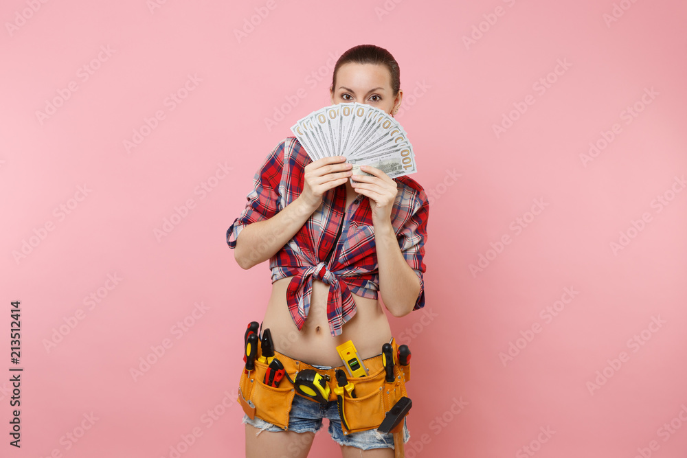 Young handyman woman in plaid shirt, denim shorts, kit tools belt full of variety instruments hold bundle of cash dollar money isolated on pink background. Female doing male work. Renovation concept.