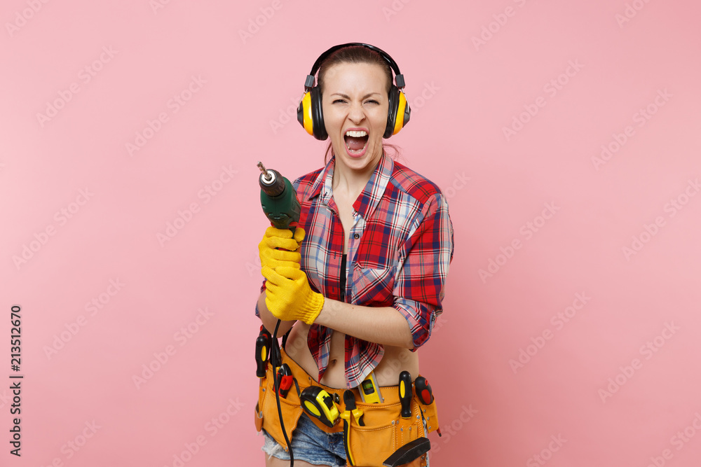 Energy sexy handyman woman in gloves, noise insulated headphones, kit tools belt full of instruments holding power electric drill isolated on pink background. Female in male work. Renovation concept. Energy sexy handyman woman in gloves, noise insulated headphones, kit tools belt full of instruments holding power electric drill isolated on pink background. Female in male work. Renovation concept.