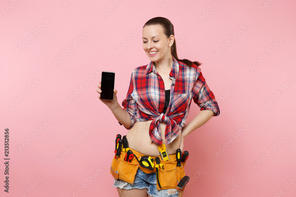 Woman with kit tools belt full of instruments holding showing camera mobile phone with blank black empty screen display touchscreen isolated on pink background. Female in male work. Renovation concept Woman with kit tools belt full of instruments holding showing camera mobile phone with blank black empty screen display touchscreen isolated on pink background. Female in male work. Renovation concept