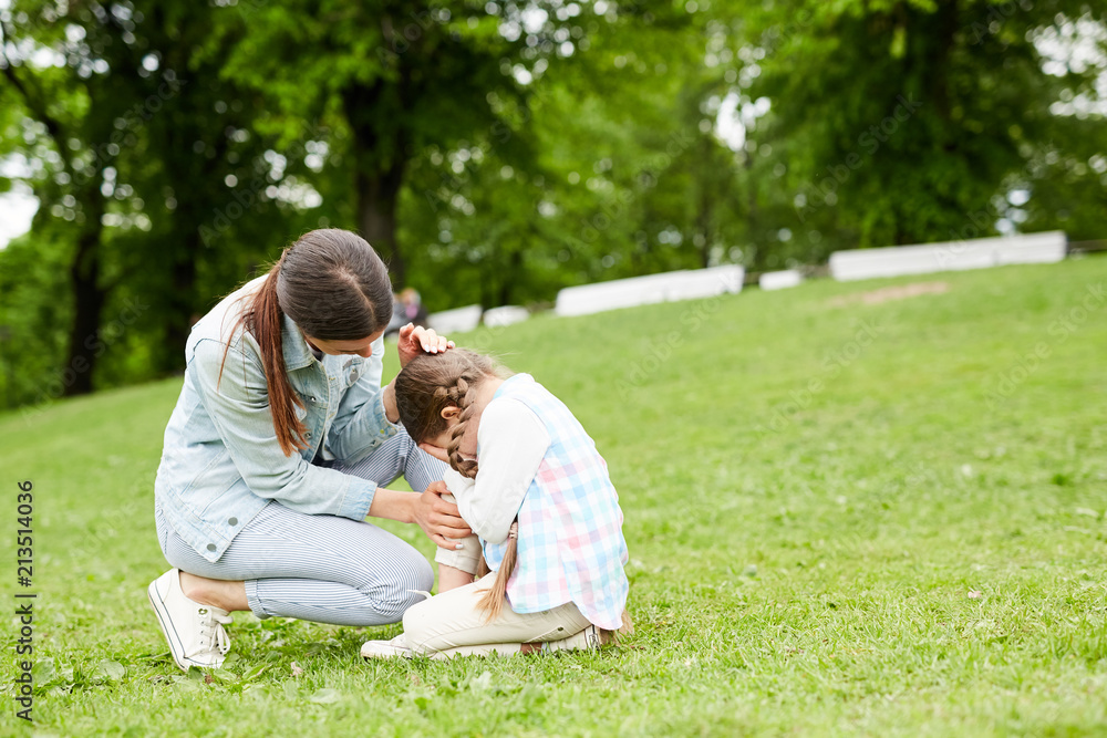 Little girl crying while sitting on green lawn and her mother comforting her Little girl crying while sitting on green lawn and her mother comforting her