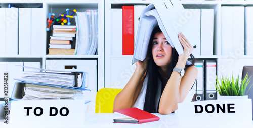 Funny overworked girl with a folder on her head sitting in the workplace cluttered with folders. Reaction of a subordinate when the boss orders her to do too much.