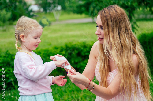 little and sweet blonde girl in a cute gray dress together with toothy smile beautiful mother wipe hands with wet wipes in the park against the background of trees and greenery