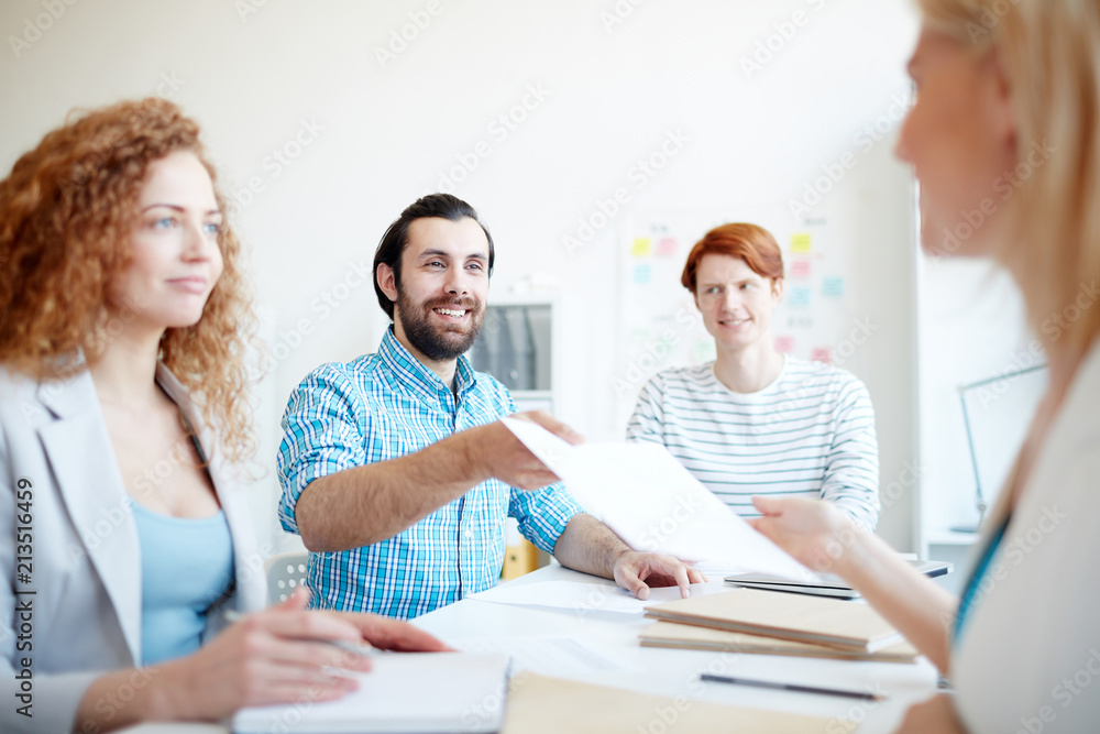 Happy young economist passing financial paper to one of colleagues or new business partner after negotiating Happy young economist passing financial paper to one of colleagues or new business partner after negotiating