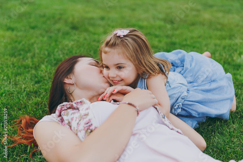 Happy woman in light dress kissing little cute child baby girl lie on green grass in park rest and have fun. Mother, little kid daughter. Mother's Day, love family, parenthood, childhood concept.