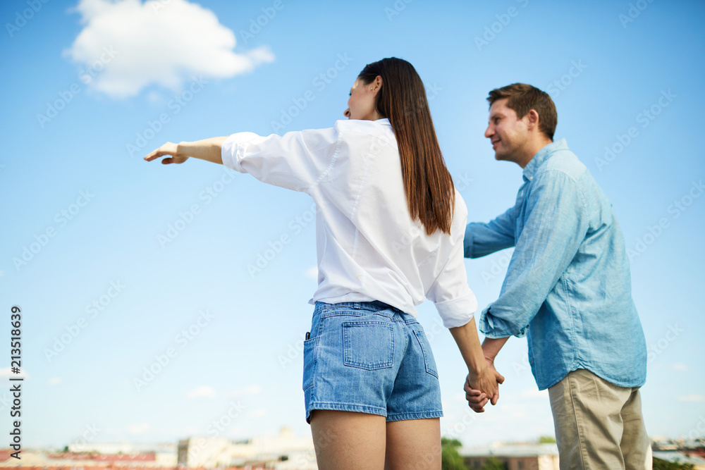 Young carefree couple in casualwear standing on roof of building and looking at cityscape Young carefree couple in casualwear standing on roof of building and looking at cityscape