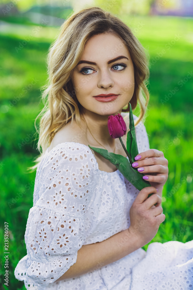 Young beautiful blonde girl in white dress with a flower tulip on a background of green grass Young beautiful blonde girl in white dress with a flower tulip on a background of green grass