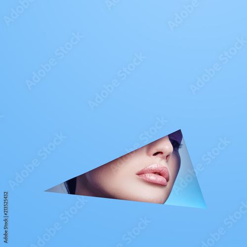 Woman looking in the hole, bright beautiful makeup, big eyes and lips, bright lipstick, professional cosmetics and facial care. Bright colored background and a gap slot in the paper © angel_nt
