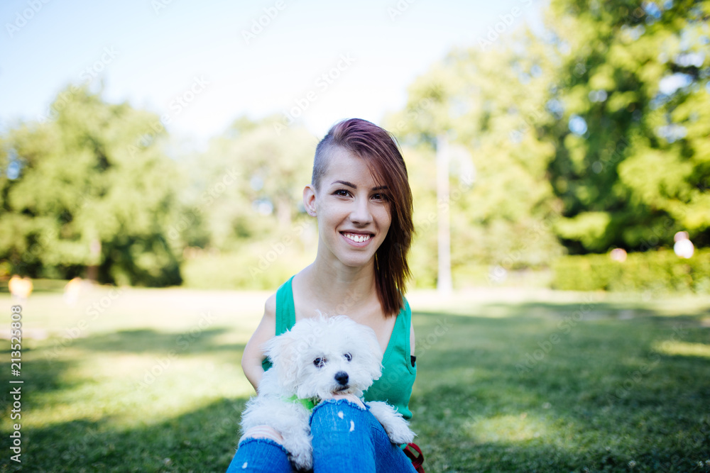 Obraz premium Portrait of beautiful smiling invalid young woman born without upper extremities enjoying with her little white puppy in park.