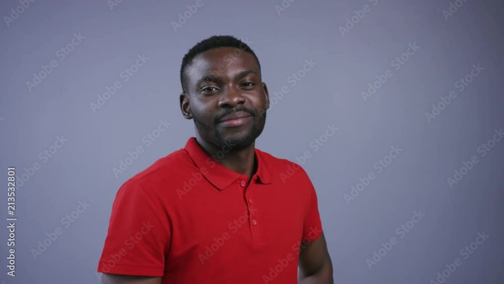 Handsome Afro-American man in bright red shirt stands still, then looks straight to camera and seductively smiles on grey background. Male beauty. Male portrait, close up view Handsome Afro-American man in bright red shirt stands still, then looks straight to camera and seductively smiles on grey background. Male beauty. Male portrait, close up view