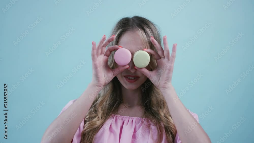 Beautiful blonde long haired woman in trendy pink dress holds delicious macaroons up to her eyes, smiles and shows her tongue on light blue background. Being a gourmet, sweets, sweet tooth. Female Beautiful blonde long haired woman in trendy pink dress holds delicious macaroons up to her eyes, smiles and shows her tongue on light blue background. Being a gourmet, sweets, sweet tooth. Female