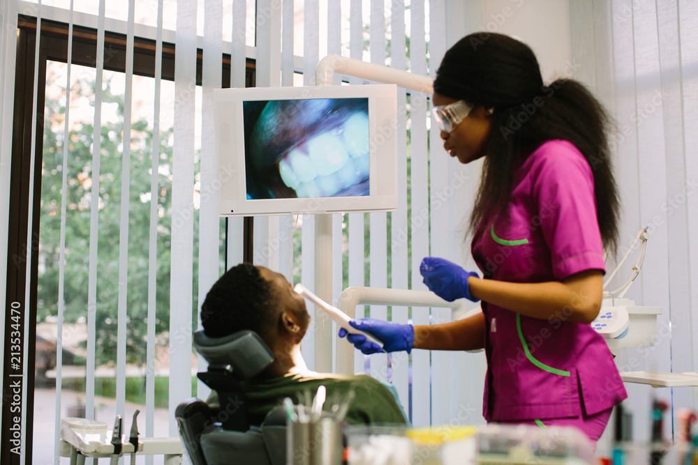 Fototapeta premium Dentisry concept, healthcare, oral and teeth care. Young beautiful african woman dentist performing diagnostics and speaking with the patient
