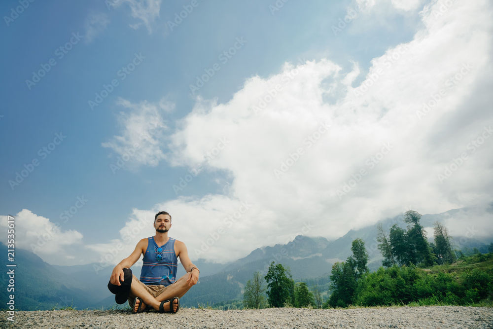 Man sits in a lotus pose and meditates against a background of a mountain and heavenly landscape.