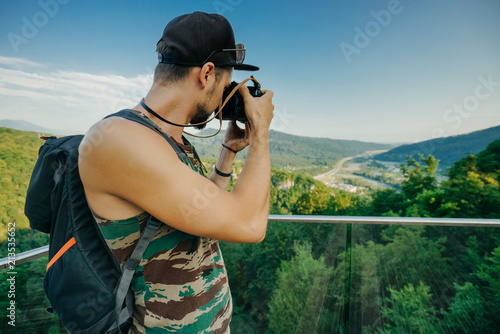 Wallpaper Mural A man photographer in a camouflage T-shirt, a cap and with a backpack holds a camera in his hands and takes a photo of a mountain landscape from a height. Torontodigital.ca