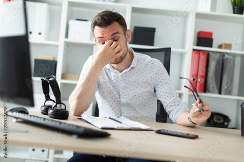 A young man sits at a table in the office, took off his glasses and rubbed his eyes.