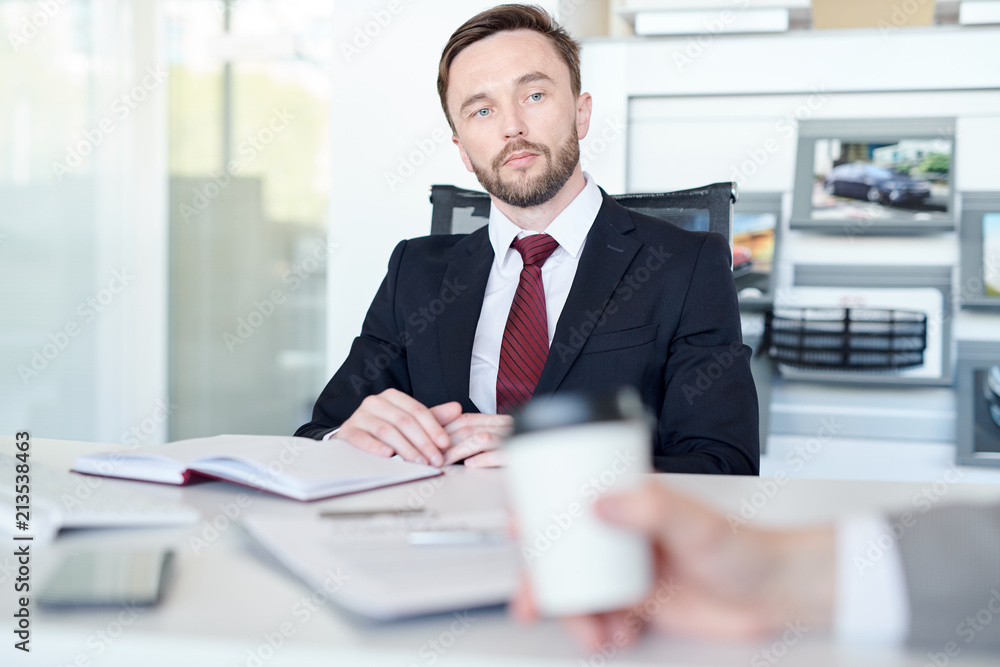 Serious young salesman sitting at desk with notebook and listening to his client Serious young salesman sitting at desk with notebook and listening to his client