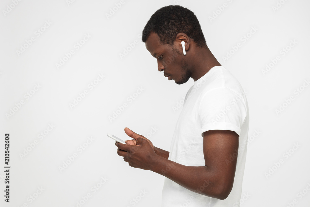 Side shot of young African man isolated on gray background holding smartphone, browsing webpages or looking for new tracks to listen through his new white wireless earphones with concerned face Side shot of young African man isolated on gray background holding smartphone, browsing webpages or looking for new tracks to listen through his new white wireless earphones with concerned face