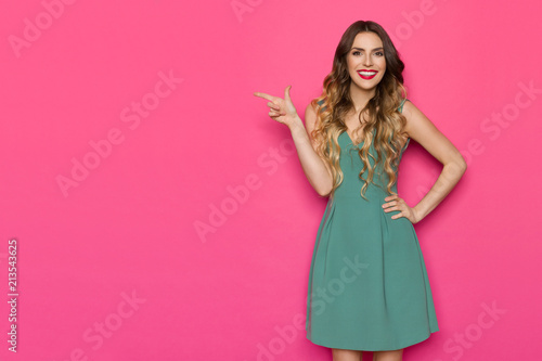 Beautiful Young Woman In Green Dress Is Pointing At Pink Copy Space And Smiling