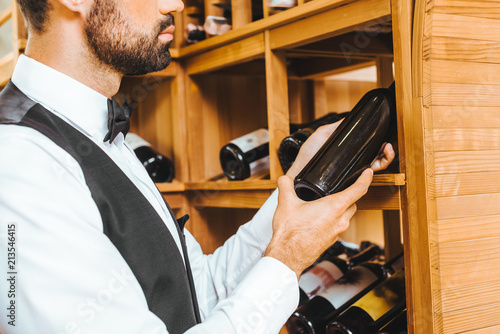 cropped shot of young wine steward taking bottle from shelf at wine store