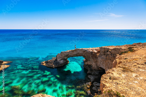 Woman on the beautiful natural rock arch near of Ayia Napa, Cavo Greco and Protaras on Cyprus island, Mediterranean Sea. Legendary bridge lovers. Amazing blue green sea and sunny day. photo