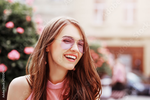 Outdoor close up portrait of young beautiful happy smiling woman wearing stylish pink aviator sunglasses posing in street of city. Copy, empty space for text