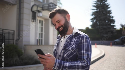 Happy smiling bearded man in fancy checkered shirt takes a walks and uses his phone, positively reacts to the news, laughs. Being online, browsing social networks, active user. Male portrait