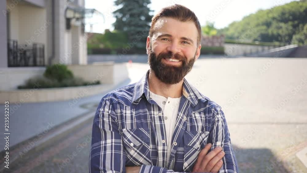 Close up view of handsome bearded man in checkered shirt waiting for someone outside, happily smiling straight to camera, crossing arms on his chest. Charisma, male power. Male portrait Close up view of handsome bearded man in checkered shirt waiting for someone outside, happily smiling straight to camera, crossing arms on his chest. Charisma, male power. Male portrait