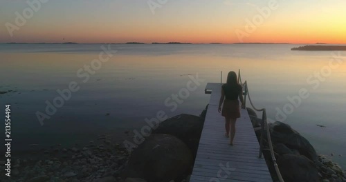 Woman on a wharf, C4K aerial view over a woman, standing at a wooden dock, enjoying the sun and a warm summer sunset, on a sunny evening dusk, in Hanko, Uusimaa, Finland
