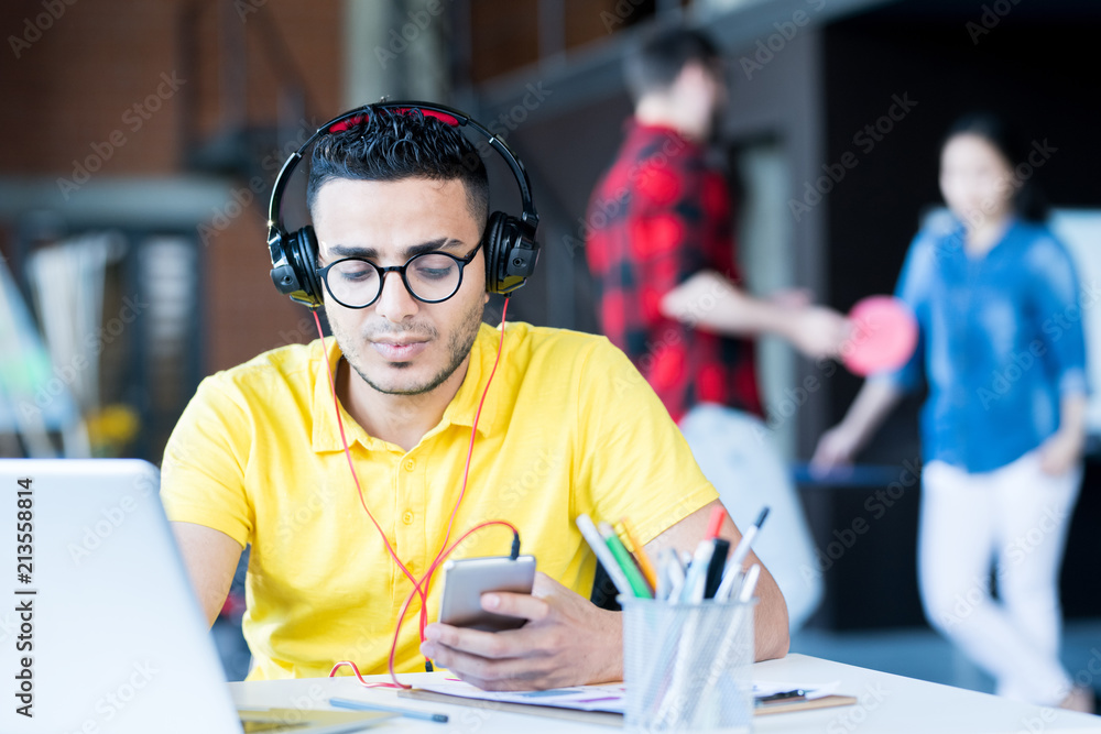 Portrait of creative young Middle-Eastern man wearing headphones and bright yellow shirt using laptop and smartphone while working in open space office of IT developers team, copy space Portrait of creative young Middle-Eastern man wearing headphones and bright yellow shirt using laptop and smartphone while working in open space office of IT developers team, copy space