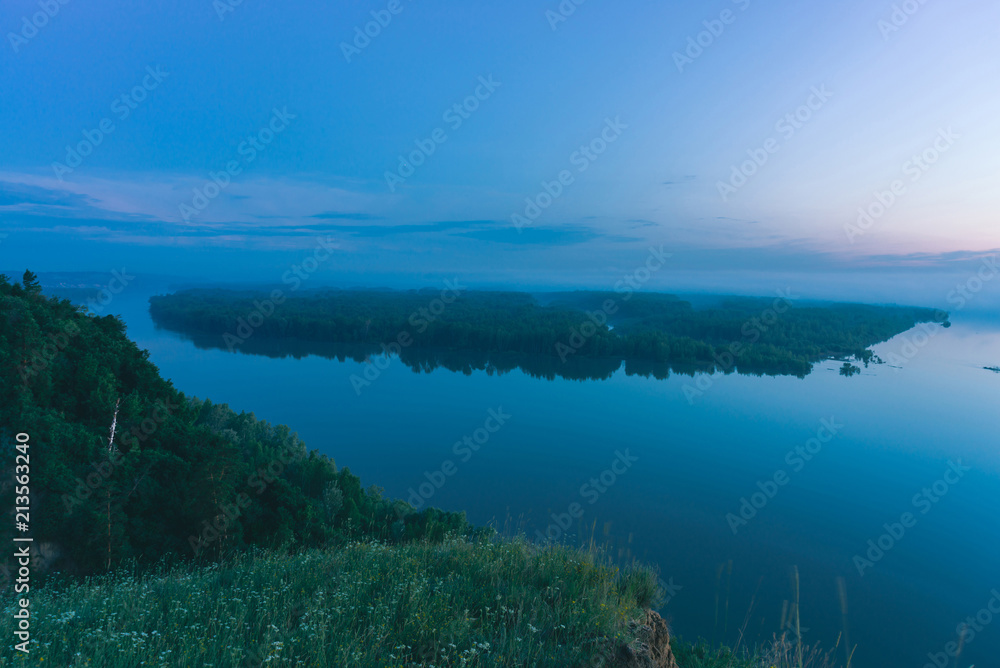 Mystical view from high shore on broad river with smooth water. Riverbank of large island with forest under mist. Early haze above trees. Morning atmospheric landscape of majestic nature in blue tone.