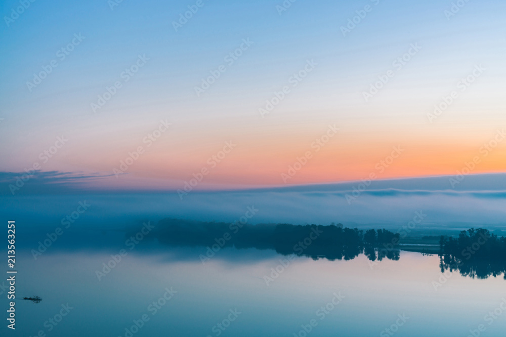 Broad river flows along diagonal shore with silhouette of forest and thick fog. Tree drifts with flow. Orange and pink glow in predawn vanila sky. Morning atmospheric landscape of majestic nature.