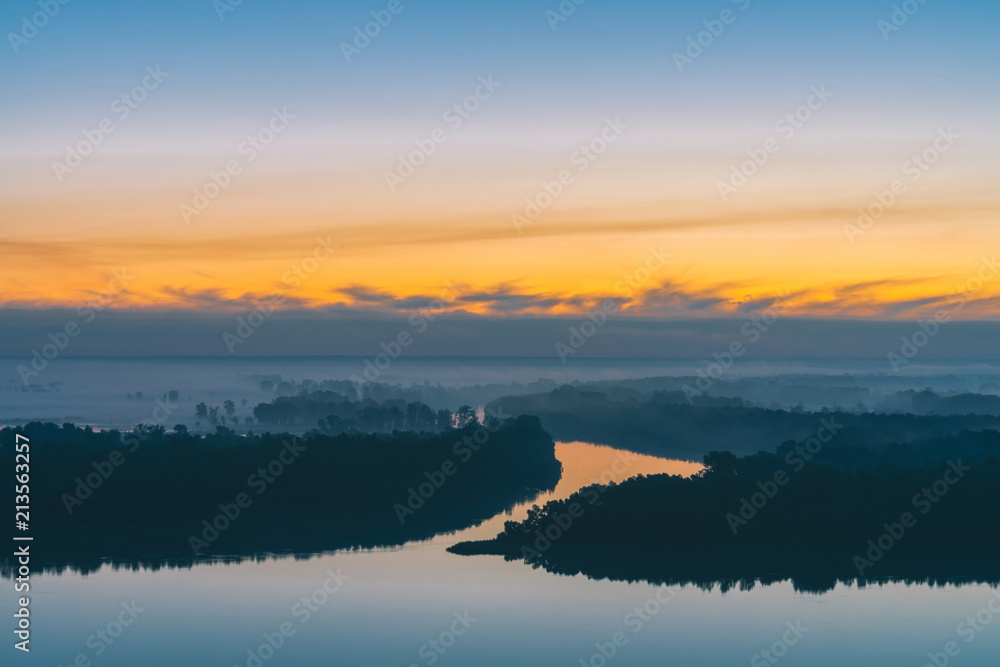 Early blue sky reflected in river water. Riverbank with forest under predawn sky. Yellow stripe in picturesque sky. Fog hid trees on island. Mystical morning atmospheric landscape of majestic nature.