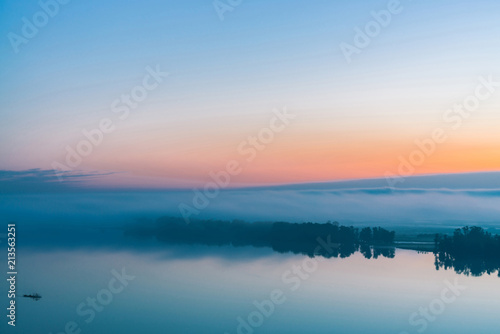 Broad river flows along diagonal shore with silhouette of forest and thick fog. Tree drifts with flow. Orange and pink glow in predawn vanila sky. Morning atmospheric landscape of majestic nature. © Daniil