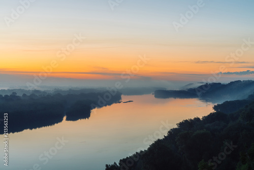 View from high shore on broad river. Riverbank with forest under thick fog. Dawn reflected in water. Yellow glow in picturesque predawn sky. Colorful morning atmospheric landscape of majestic nature.