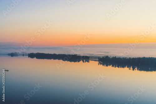 Fototapeta Naklejka Na Ścianę i Meble -  Broad mystical river flows along diagonal shore with silhouette of trees and thick fog. Gold glow in predawn sky. Calm morning atmospheric landscape of majestic nature in warm tones.